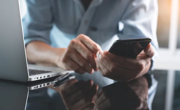 Person in front of a laptop looking at mobile phone.